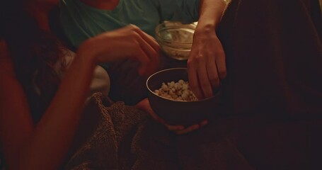 A young couple shares popcorn and laughs while watching a movie at home on the couch. They're under a blanket and enjoying the evening. - Powered by Adobe