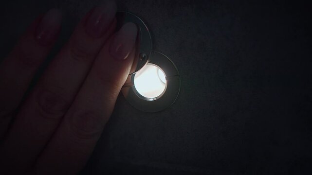 A close-up of a door peephole, in the darkness, a woman's fingers are pushing back the flap, there is a bright light behind the door.