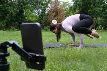 A woman is practicing yoga outdoors, following an online session on her phone. She sits on the grass in a peaceful natural setting, focused on the screen in front of her. 