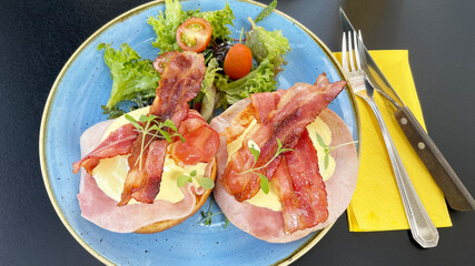 eggs benedict served with crispy bacon, ham and hollandaise sauce on brioche bun and side salad for breakfast or brunch