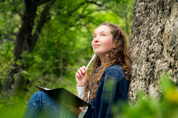 Portrait of young woman with curly hair holding pen to chin looking up and dreaming writing in notebook relaxing resting alone dressed in blue shirt and jeans sitting near rock in forest outdoors