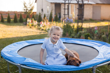 a three year old smiling girl playing with dachshund dog on trampoline in green sunny summer park