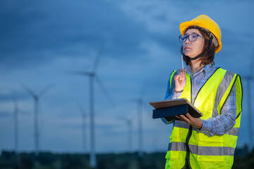 A woman technician wearing a yellow vest and a hard hat points to a tablet. She is standing in front of a field of wind turbines