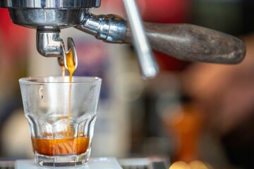 Close-up of espresso being poured from a coffee machine spout into a clear glass cup with a blurred background, highlighting freshness and rich coffee flavor