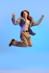 Obraz premium Teen girl jumping in air with joyful face and arms raised in casual clothes against blue background in vivid neon light. Success. Concept of excitement, freedom, happiness, youth energy, emotion.