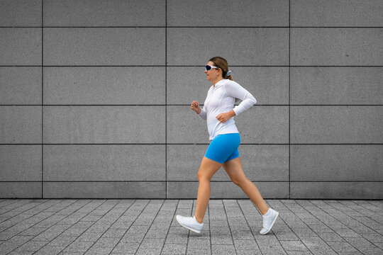 Beautiful middle aged woman running against background of concrete slabs in city on summer day. Side view
