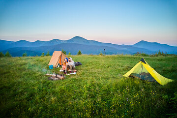 Scenic mountain campsite with two tents in morning. Two campers enjoying serene view of rolling mountains under clear sky. Picturesque sunrise landscape, creating tranquil and cozy outdoor experience.