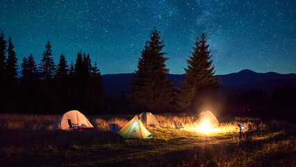 Several tents around glowing campfire on grassy clearing under starry sky. Warm light of campfire illuminates scene, creating cozy and serene night camping atmosphere in the mountains.