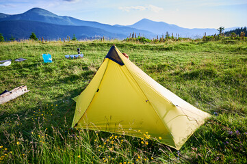 Yellow tourist tent on grassy hillside with camping gear scattered nearby. Stunning view of green hills, distant mountains under clear blue sky, creating perfect spot for peaceful outdoor adventure.