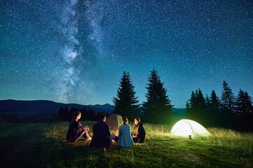 Group of friends hikers resting around campfire, surrounded by illuminated tents under stunning starry sky. Milky Way stretches above, creating magical atmosphere in peaceful mountain wilderness.