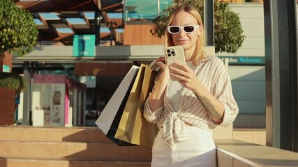 Blonde woman carrying shopping bags, browsing smartphone with sunglasses, standing in colorful downtown setting during sunny shopping day.Consumerism, sale, black friday. Online shopping.