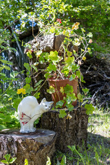 Flower pot with green plant on tree stump and white ceramic cat sculpture in sunny garden.