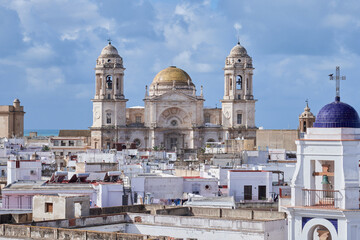 Panoramic view of the Cathedral of C&aacute;diz, Spain, captured from the rooftops of the old town. The iconic twin towers and golden dome rise above the dense whitewashed buildings under a partly cloudy sky