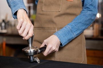 Close-up of young Asian barista wearing brown apron and denim shirt tamping ground coffee into portafilter. Professional hands pressing coffee evenly on black mat, preparing for espresso extraction 