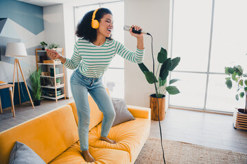 Cheerful woman singing on a vibrant couch in a sunny modern living room, expressing joy and leisure...
