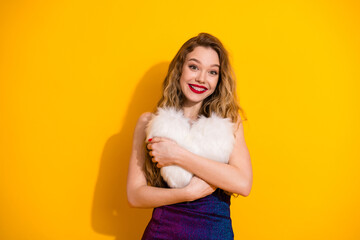 Cheerful young woman in fashionable dress holding a soft heart pillow against a bright yellow...