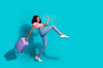 Young woman holding a suitcase on a turquoise background, expressing excitement and displaying a...