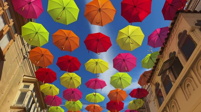 Bright multicolored umbrellas suspended over a street with traditional Mediterranean architecture under a radiant blue sky. A cheerful urban installation creates a festive atmosphere.