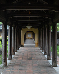 Outdoor long passage with round wooden columns on the sides. An open archway in the distance. 