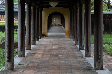 Outdoor long passage with round wooden columns on the sides. An open archway in the distance. 