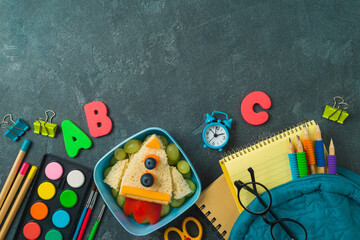 Back to school concept with healthy lunch box, sandwich rocket, backpack and school supplies on dark background. Top view, flat lay