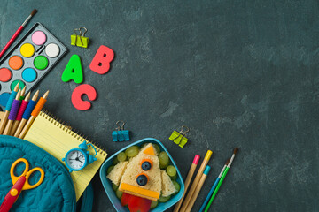 Back to school concept with healthy lunch box, sandwich rocket, backpack and school supplies on dark background. Top view, flat lay