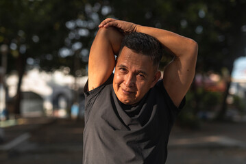 A middle aged man smiling while doing a tricep stretch, preparing for exercising at the park....