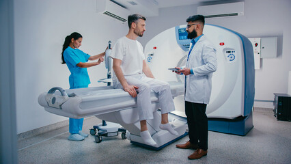 Attractive male sitting upright on MRI bed having discussion with doctor holding digital tablet. Smiling nurse adjusting nearby device while preparing for procedure in clean clinical environment.