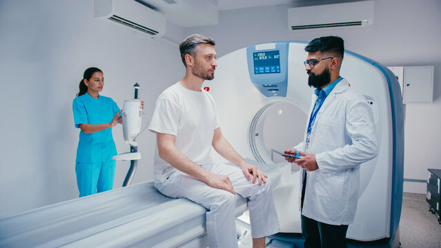 Man describing lower back pain while seated on exam table next to MRI scanner to his doctor. Bearded practitioner reviewing notes on tablet as nurse in blue uniform adjusting equipment in background.