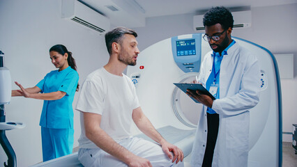 Man seated on examination bed describing condition while African American doctor writing on clipboard. Female nurse in turquoise scrubs preparing imaging device in well-lit hospital room.