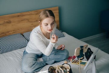 Teenager applying makeup while recording video tutorial on bed