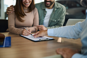 Unrecognizable young couple signing a contract on a meeting with salesperson in a car showroom.