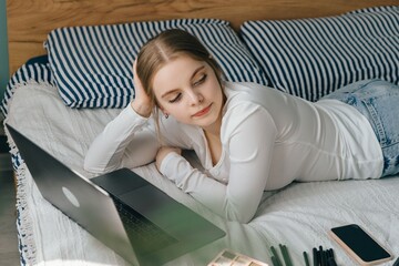 Teenage girl relaxing on bed using laptop for online learning and makeup tools