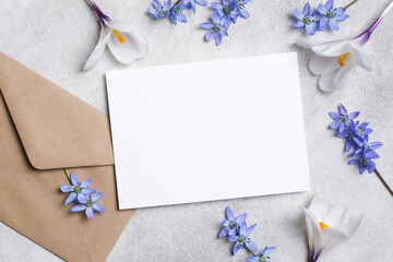 Blank invitation card mockup with kraft envelope and blue spring flowers on light concrete surface, flat lay for wedding or greeting cards