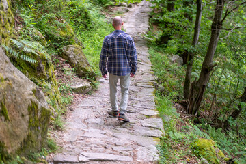 Obraz premium Young traveler man walks on old stone path through serene green forest, Italian Nature Hike, Exploring Ancient Trails, Hiker on historic pathway surrounded, Rural wilderness Journey