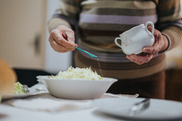 Close-up image of an elderly woman cooking at home, sprinkling seasoning onto a fresh green salad in a white bowl.