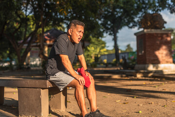Unfit Southeast Asian man in his 50s sits on a bench, catching his breath after an intense jog or...
