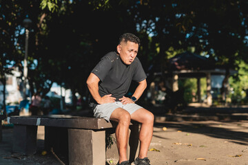 Middle aged Southeast Asian man holds stomach in discomfort while seated on park bench, possibly...