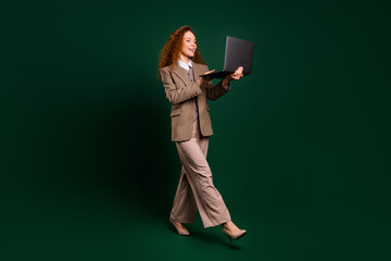 Confident businesswoman in formal attire holding a laptop, walking confidently against an emerald...