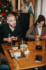 A group of individuals seated at a dining table, interacting in a warm festive ambiance featuring a decorated Christmas tree. They enjoy beverages, food, and discussions in a cozy setting.