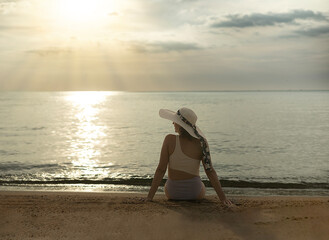 A woman sits sunbathing on the beach. The waves gently crash against the shore. Looking at the sun makes her feel peaceful.