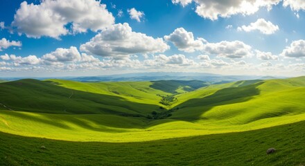 Obraz premium Panoramic view of rolling green hills and valleys under a vibrant blue sky dotted with fluffy white clouds. Sunlight casts shadows, enhancing the landscape's texture and depth