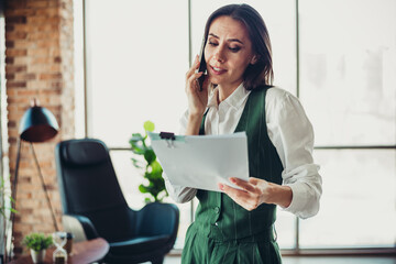 Plakat Confident businesswoman talks on phone while reviewing documents in stylish modern office environment