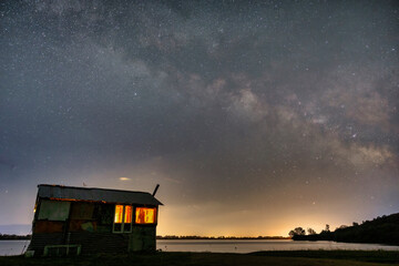 A tranquil night at Lake Kerkini, where a wooden cabin glows warmly under a breathtaking Milky Way
