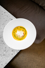 A bowl of creamy pumpkin soup garnished with shredded cheese and green herb oil, served on a white plate on a speckled gray countertop. The soup’s vibrant colors make it visually appealing