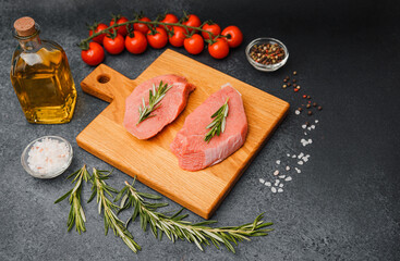 Raw veal steaks with rosemary on cutting board surrounded by cherry tomatoes oil salt and pepper..