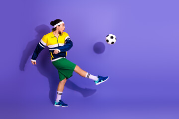 Young man in retro activewear playing soccer against a violet background showcasing sporty...