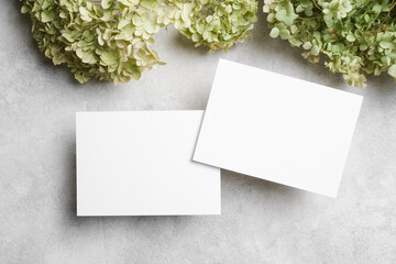 Two blank horizontal greeting cards mockup on light textured background, surrounded by soft green hydrangeas, ideal for stationery or invitation design showcase