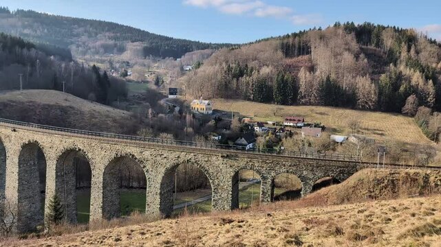 A bridge with a train track winds past a small village surrounded by thick woods in a rural area during fall