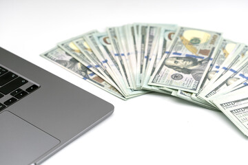 Stack of hundred dollar bills is fanned out next to laptop, symbolizing online income and financial...
